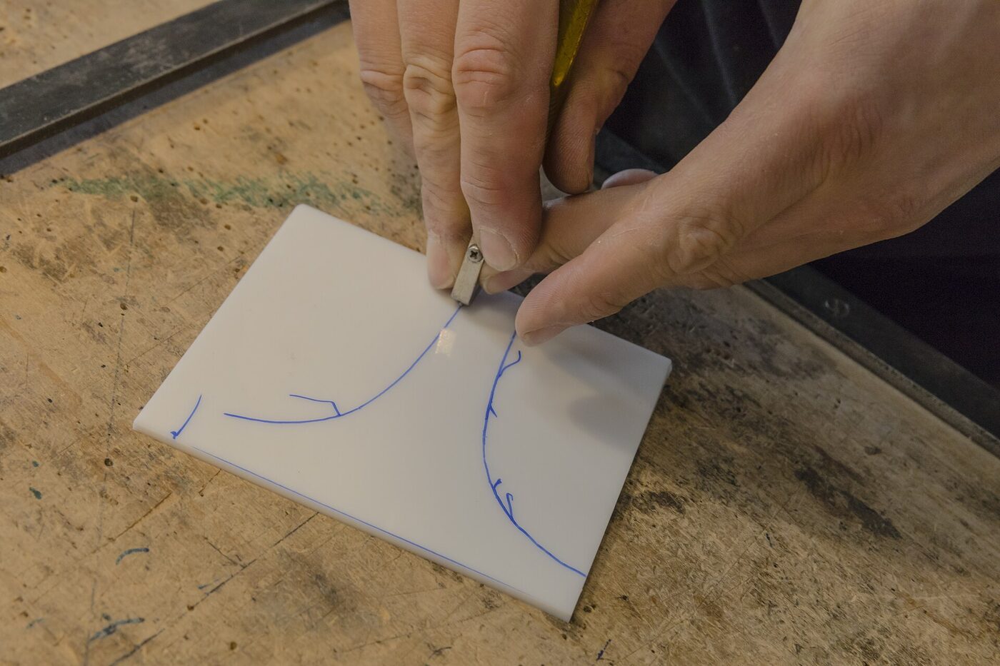 A glazier's hands scoring a sheet of white glass along a curved line drawn in blue ink, using a small hand cutter on a workbench