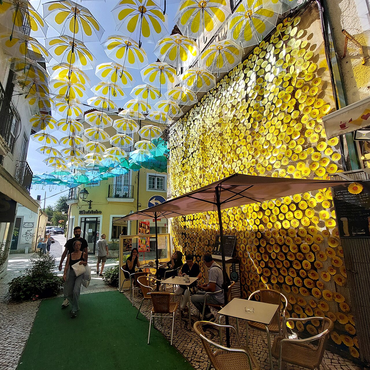 A café terrace in Águeda under a canopy of yellow and blue parasols, beside a wall covered in a shimmering yellow installation