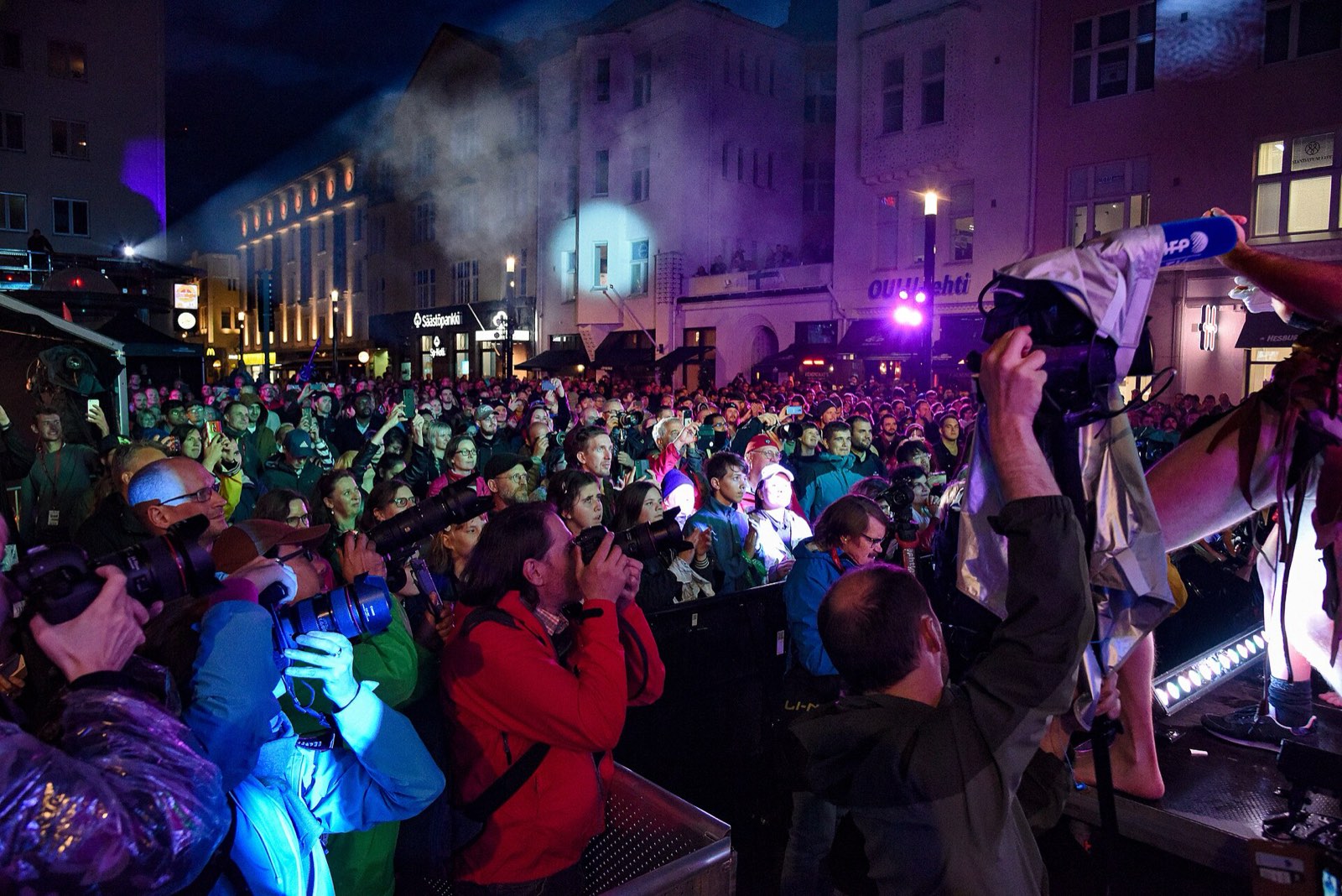 A dense crowd at night pressed against the stage barrier in Oulu, photographers with long lenses and a news microphone raised toward a performer at the edge of frame