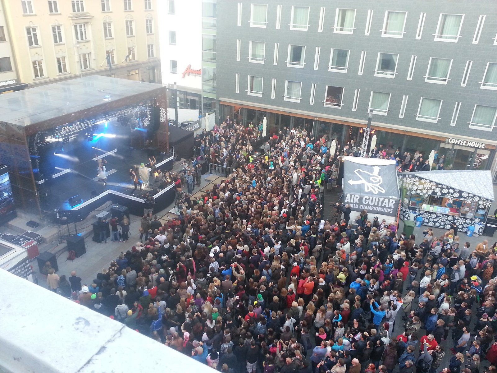 Aerial view of a packed city square in Oulu, thousands of spectators facing a lit stage, with a large banner reading AIR GUITAR WORLD CHAMPIONSHIPS at the right