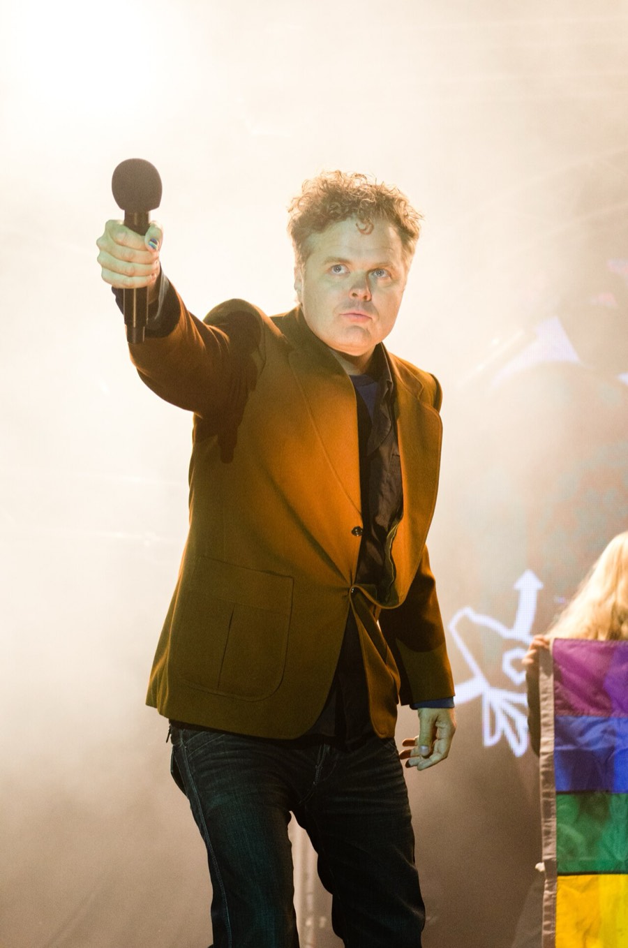 Jukka Takalo on stage in an orange blazer, holding a microphone out toward the audience, backlit by warm stage light