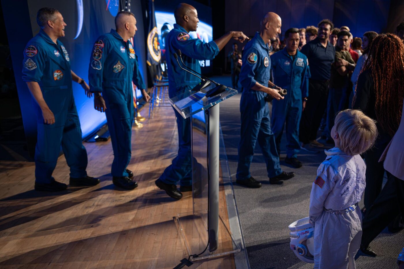 A small child in a white astronaut costume, holding a toy helmet under one arm, stands at the edge of a stage watching five astronauts in blue flight suits