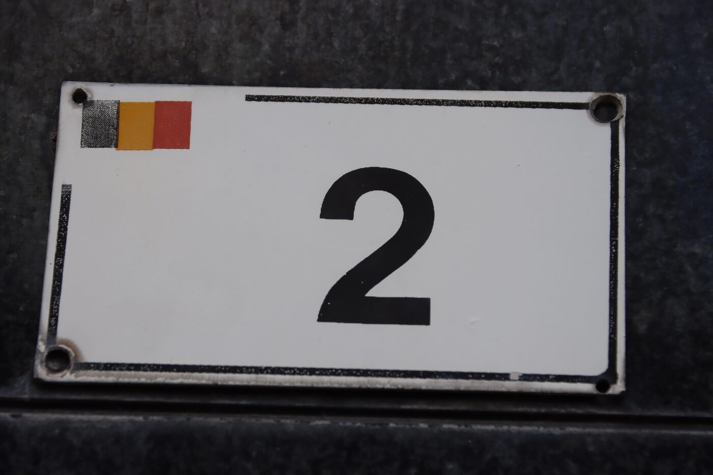 An enamel house-number plate reading 2 with a small Belgian flag in black, yellow and red printed in the top-left corner