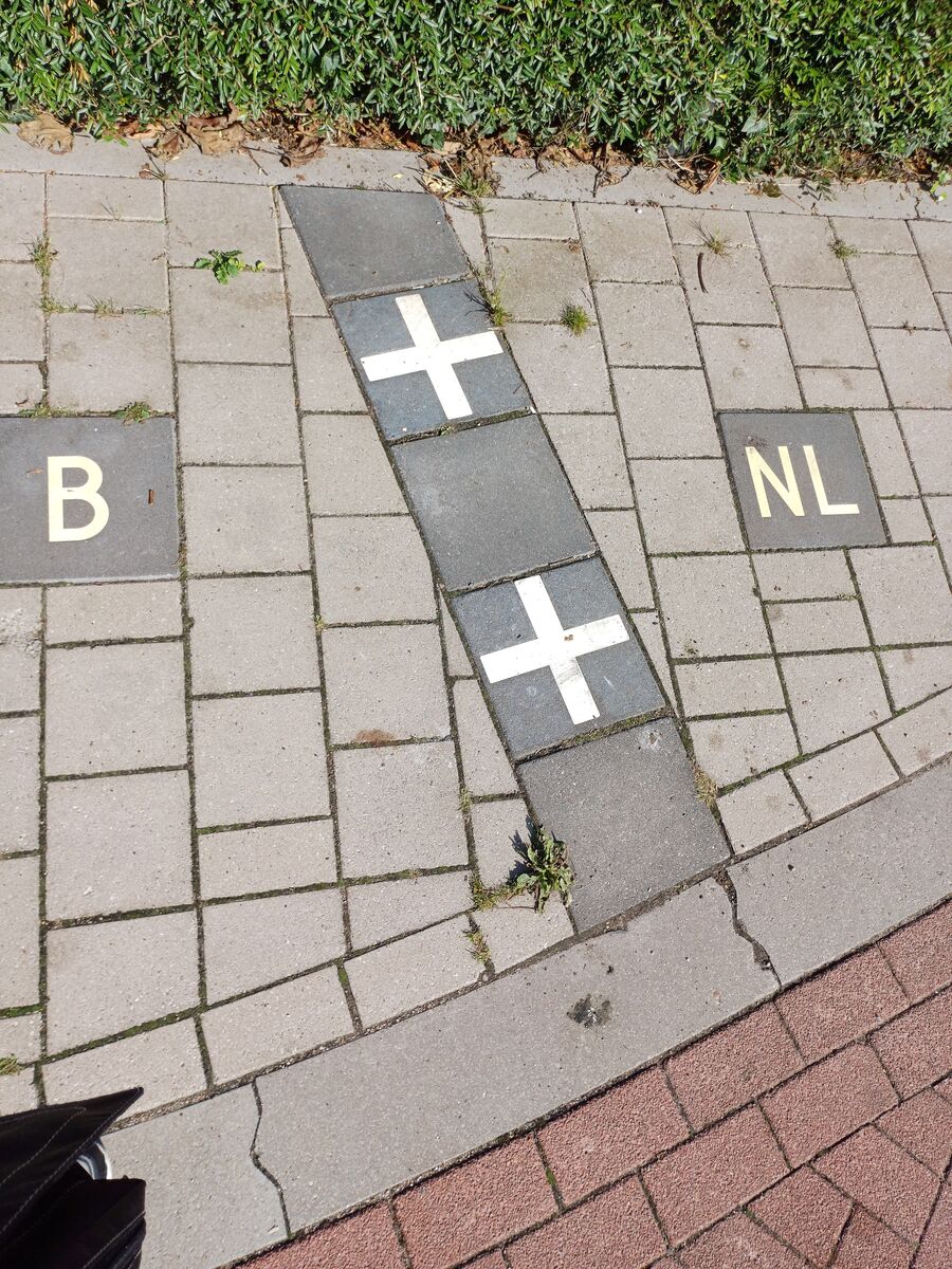 A pavement in Baarle with a line of dark tiles bearing white crosses running diagonally across it, a tile lettered B on one side and a tile lettered NL on the other