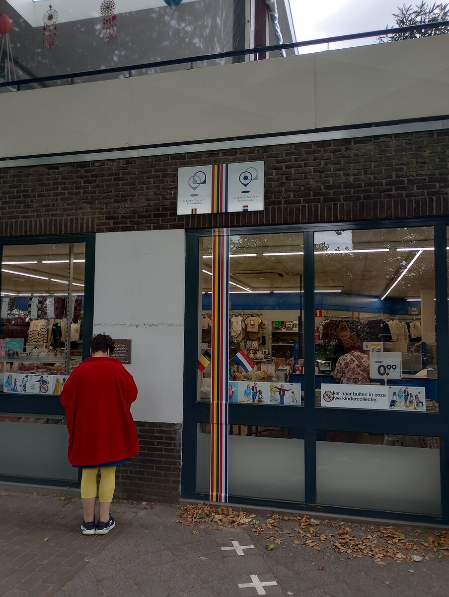 A shop front in Baarle with a striped border line painted up the wall and across the window, signs above reading you are in Baarle-Hertog and you are in Baarle-Nassau, and white crosses in the pavement below