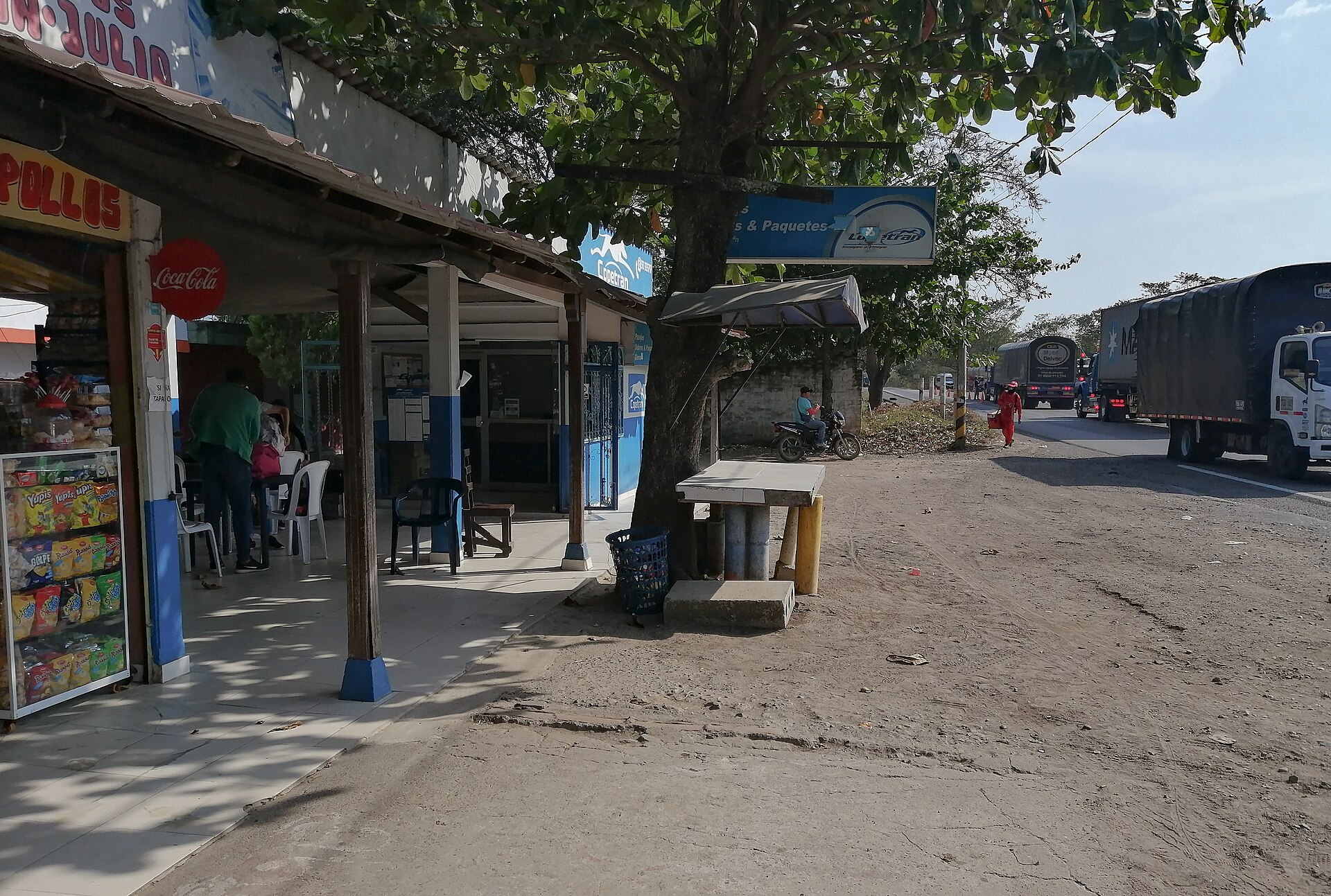 A dusty roadside row of small shops with metal awnings beside a highway where cargo trucks pass, in a rural Colombian town