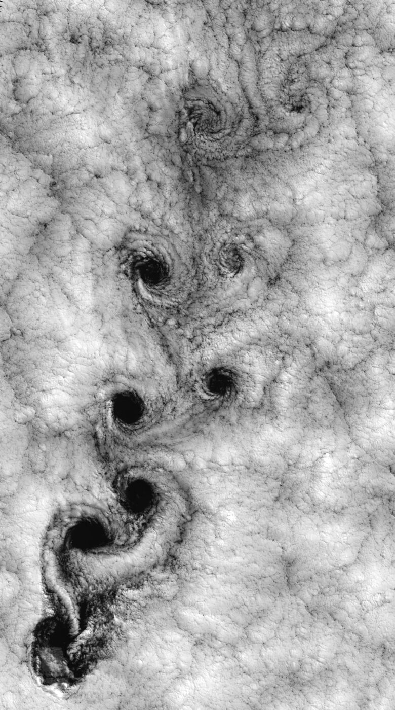 Black and white satellite image of a thick blanket of low cloud, torn into a long descending chain of dark spirals — one curling clockwise, the next counter-clockwise — running from the bottom of the frame toward the top
