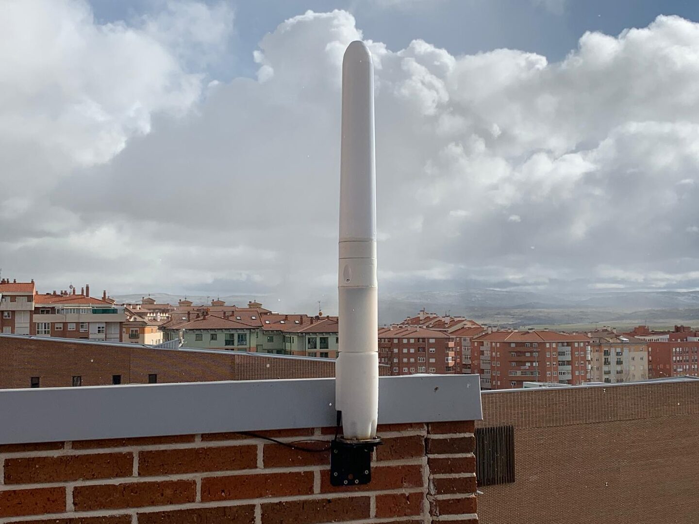 A small white bladeless Vortex mast bolted to a black bracket on a red brick parapet at the edge of a flat roof, with the tiled roofs and apartment blocks of a Spanish town and low hills stretching away under a cloudy sky behind it