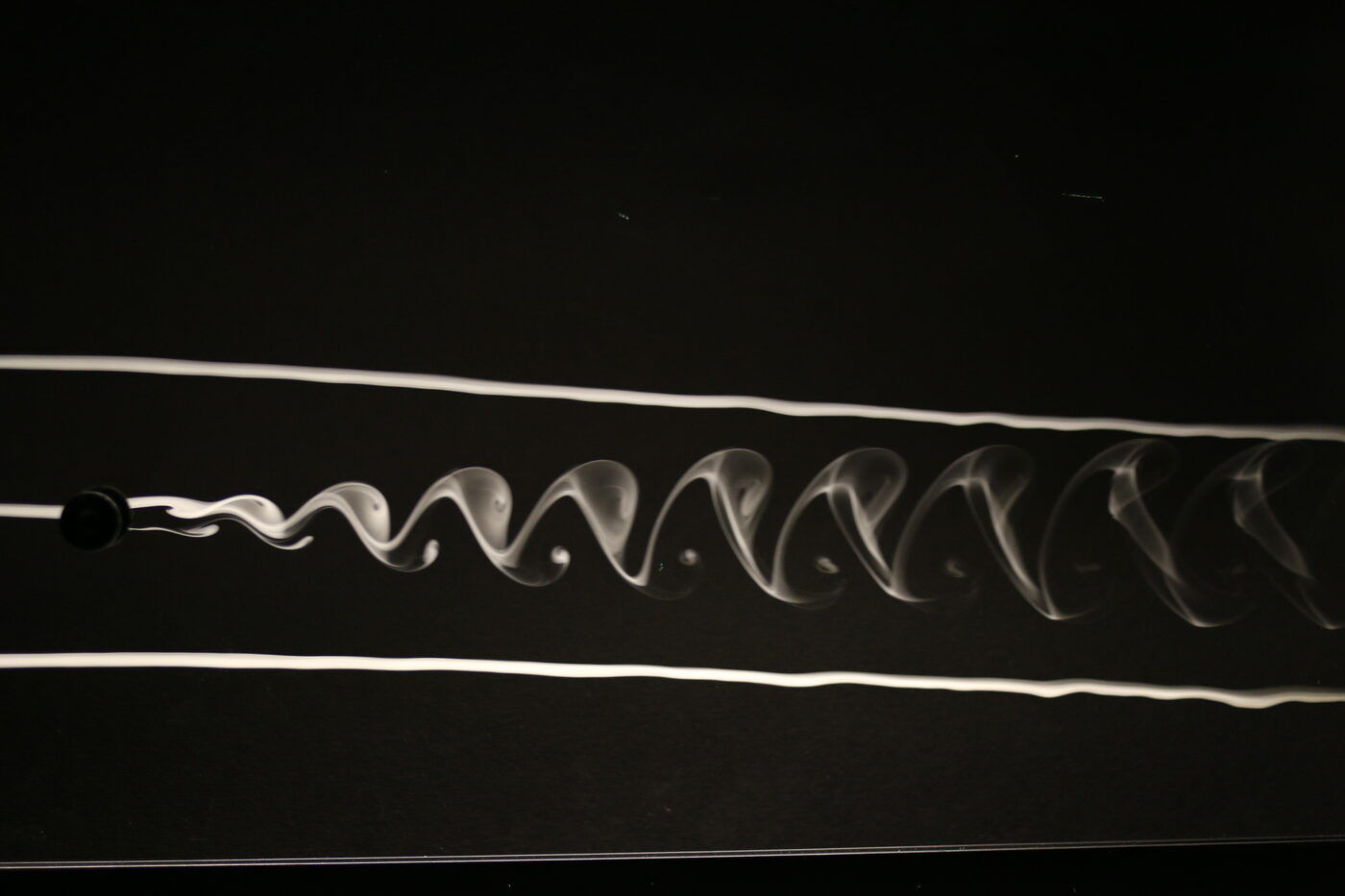 Laboratory photograph on a black background: a stream of white smoke flows from left to right past a small black cylinder, and immediately behind the cylinder the smooth smoke line breaks into a long chain of alternating curls, one above, one below, repeating down the frame