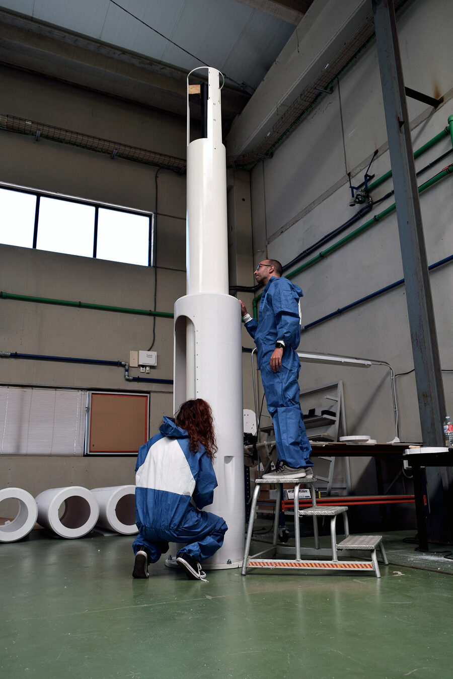 Two engineers in blue coveralls assemble a tall white cylindrical mast in a workshop — one crouches at the base while the other stands on a step stool to reach the top, where a dark inner rod is visible inside the open end of the white outer shell; spare white cylinder sections lie on the floor behind them