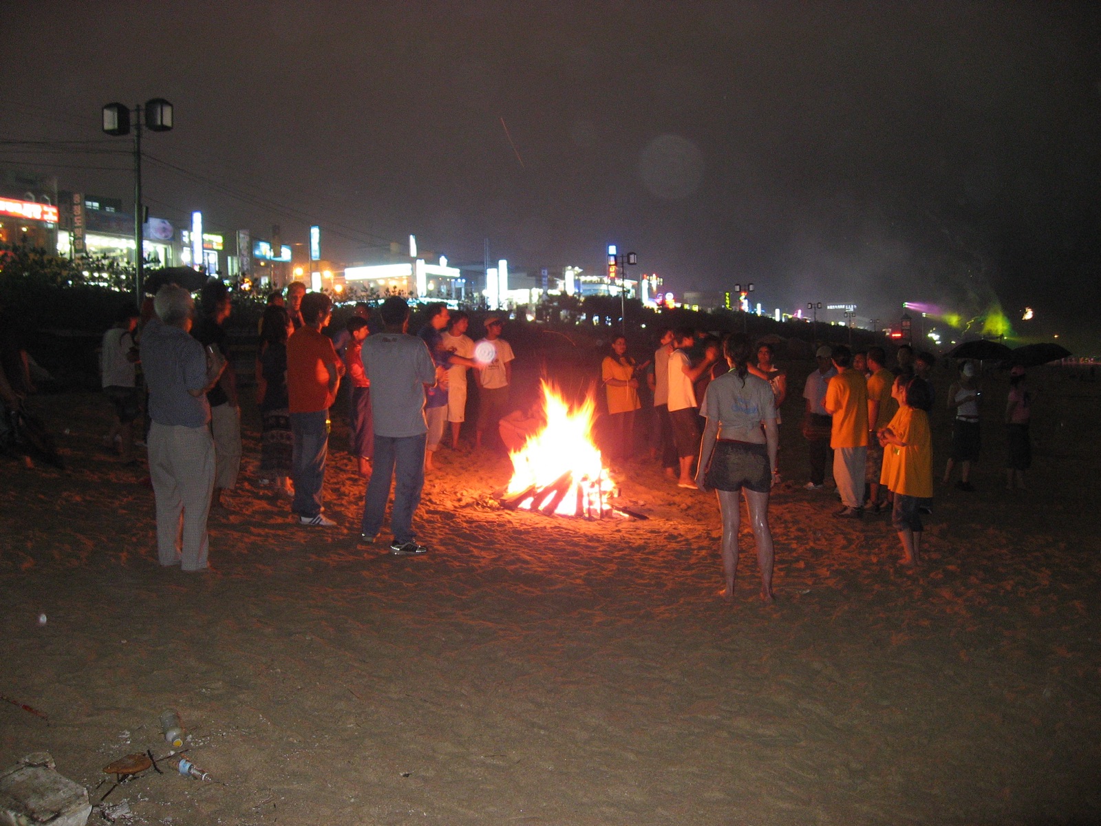 A crowd gathered around a beach bonfire at night during the Boryeong Mud Festival, with the lit-up seafront behind them