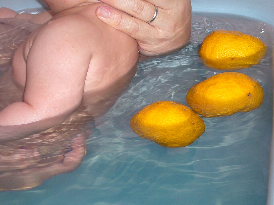 A person's arm and shoulder in a hot bath with three whole yellow yuzu fruits floating on the water beside them