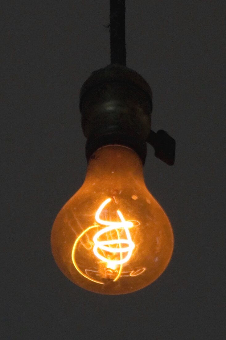 Close-up of the Centennial Light: a pear-shaped bulb glowing dim orange on an old brass socket, hanging in a dark room