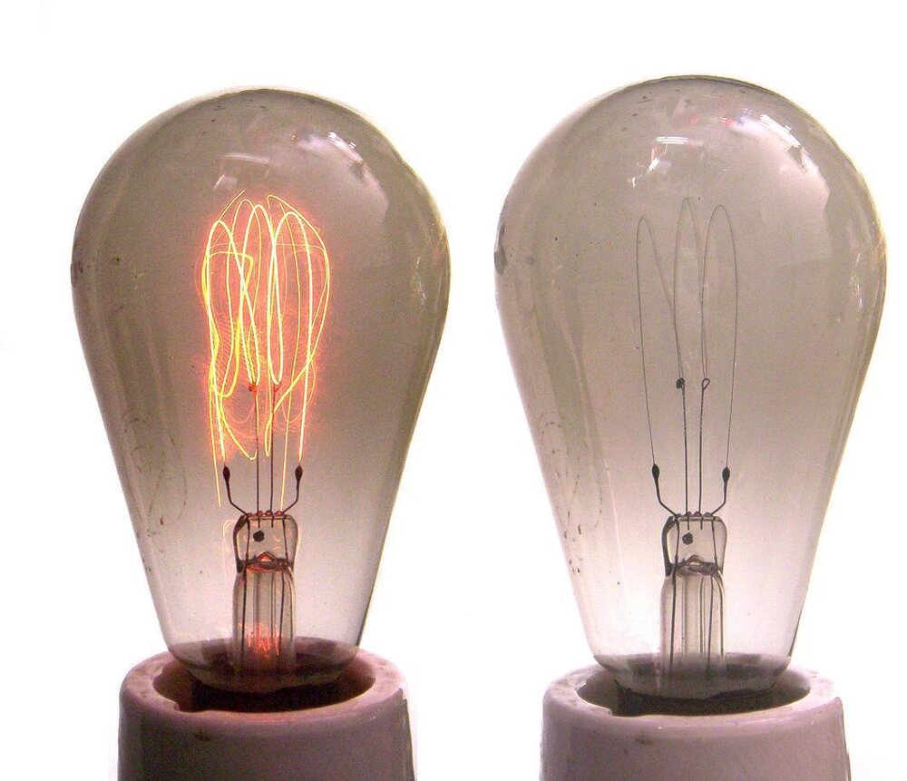 Two pear-shaped carbon-filament bulbs side by side, the left one lit with a dim orange hairpin filament, the right one unlit showing the dark filament and a grey haze on the glass