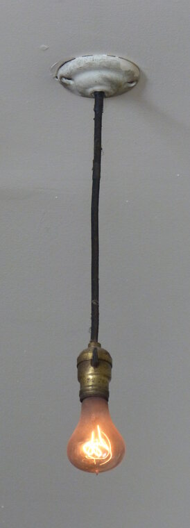 The Centennial Light hanging on a long black cord from a plain painted ceiling plate, with a brass socket and the small glowing bulb at the bottom