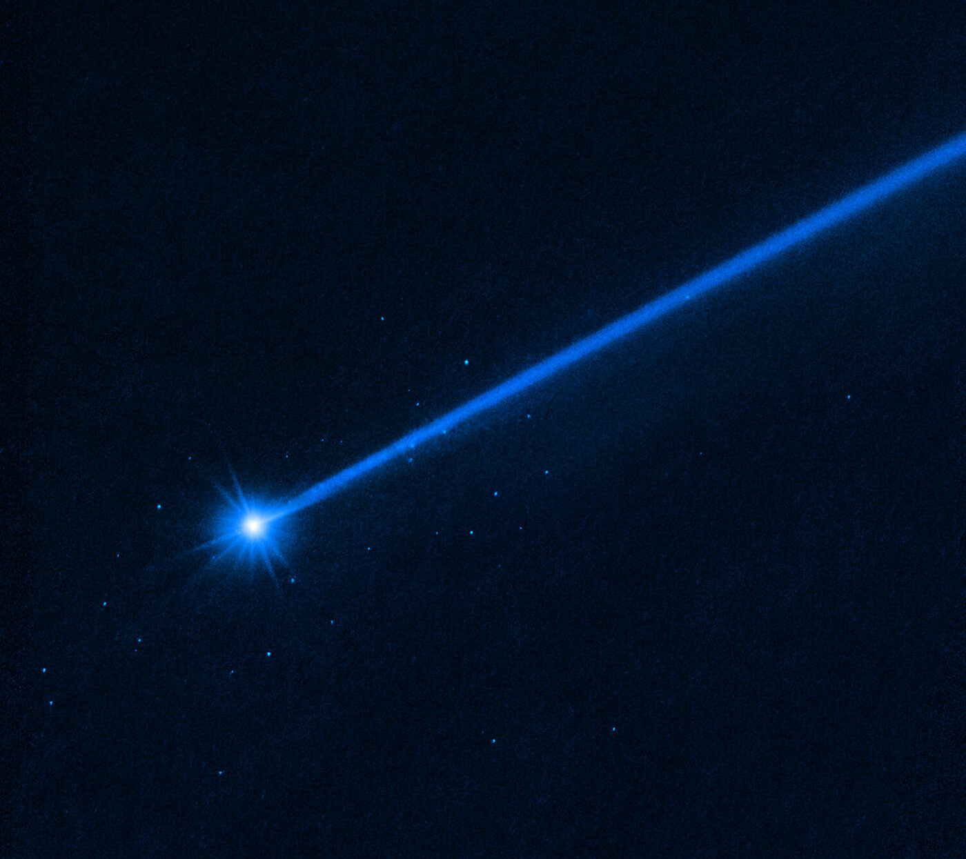 Deep blue Hubble image with a brilliant white starburst at lower left and a straight bright dust tail streaming to the upper right, scattered with faint pinpoints of light