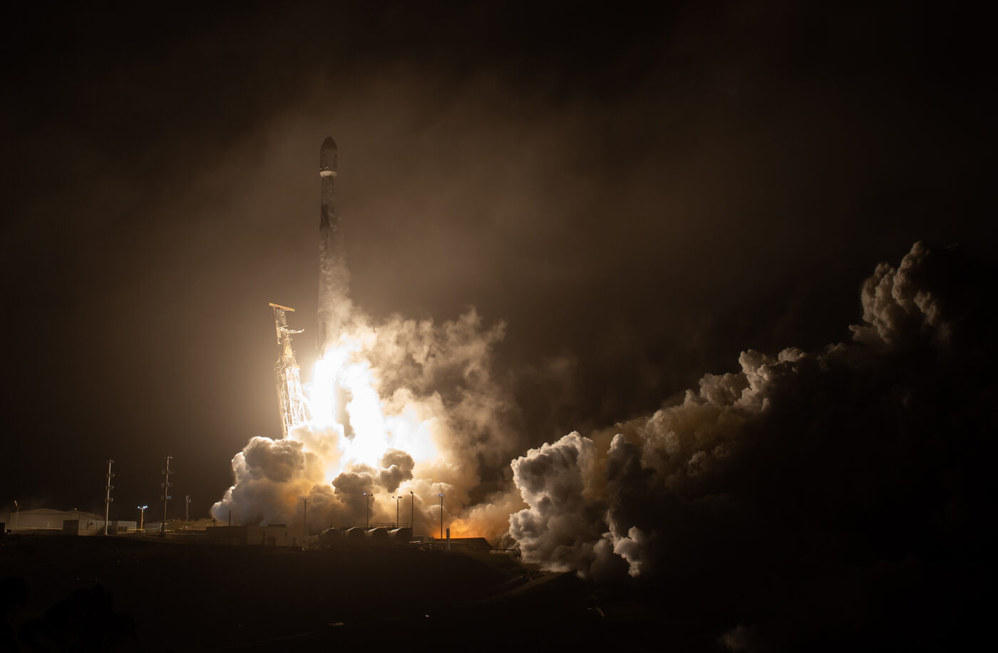 A Falcon 9 rocket lifting off at night, bathed in its own white-gold engine light, with billowing smoke rolling across a dark launch complex