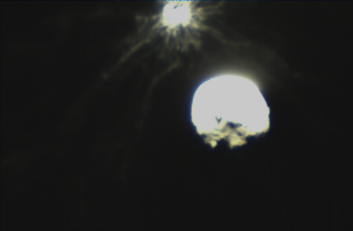 Grainy dark image showing two bright blobs: a small brilliant point at top and a larger glowing lumpy mass below, surrounded by faint wispy streamers of debris fanning outward