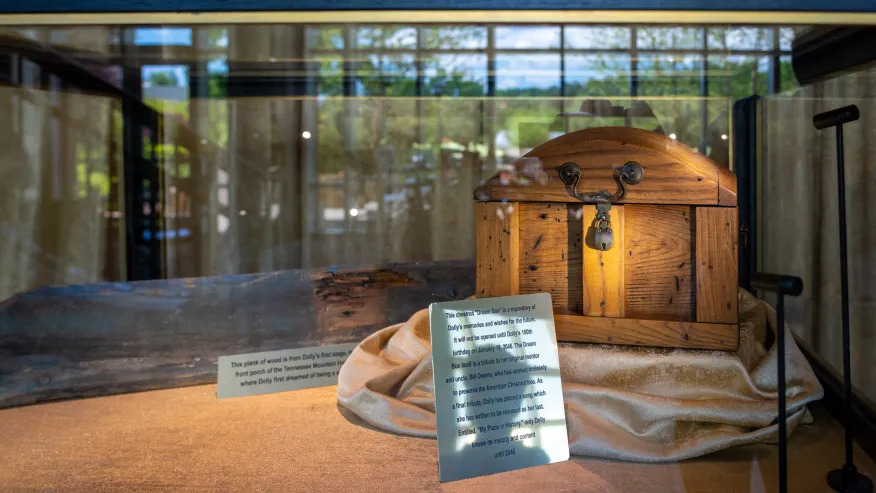 A small wooden chest with an arched lid and a brass padlock sits on a rumpled fold of pale cream velvet inside a glass display case. To its left, a long weathered plank of dark grey wood lies flat on the case floor. Two engraved placards lean upright in front, one beside the plank and a larger one against the velvet. The glass carries reflections of the resort's windows and the trees outside.