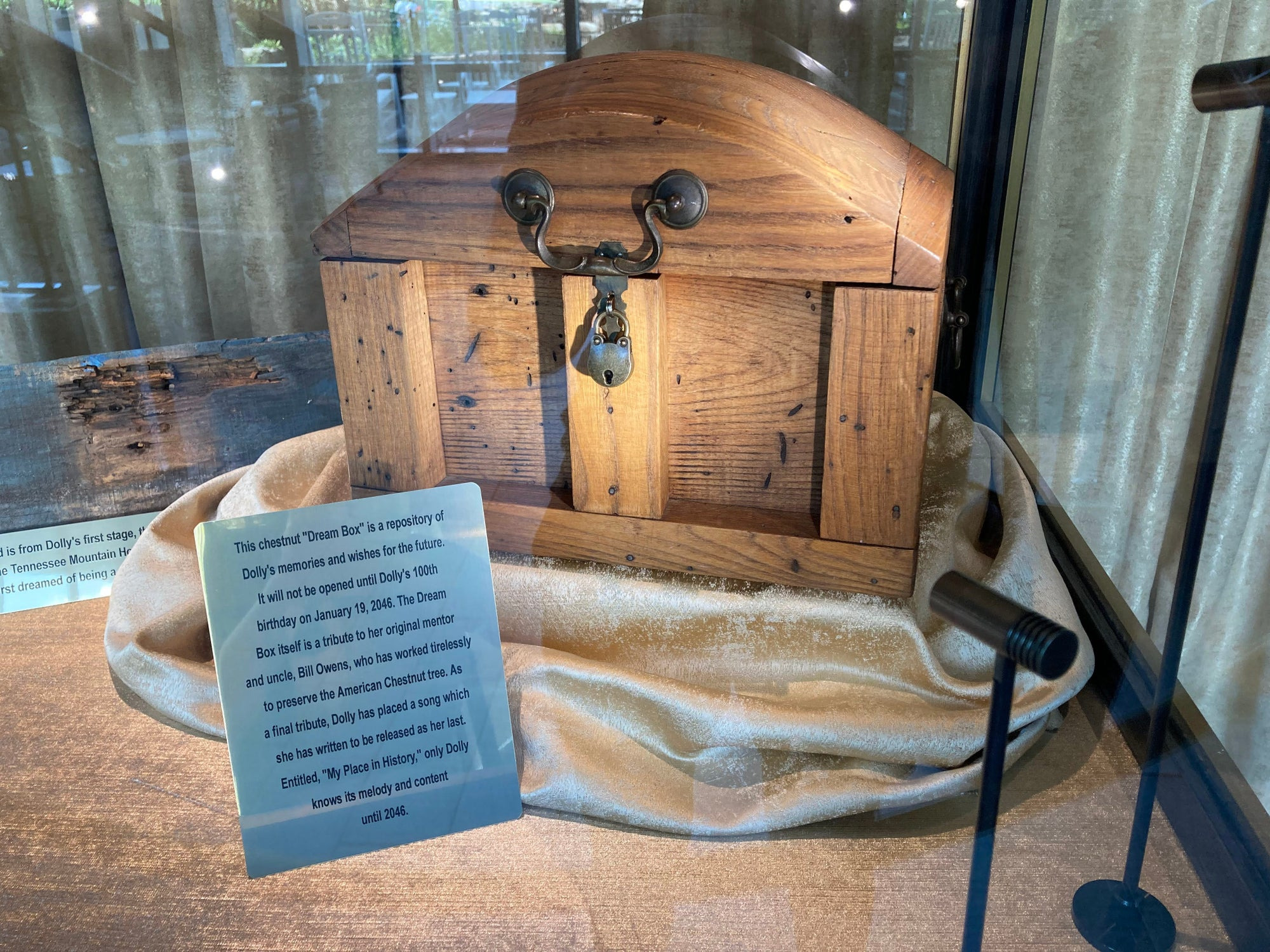 A close view through display glass of a chestnut chest with an arched lid, a curved metal bail handle mounted across the front of the lid, and a brass padlock hanging from the clasp beneath it. A printed placard stands upright on the velvet in front of the chest, its lines of text running edge to edge.