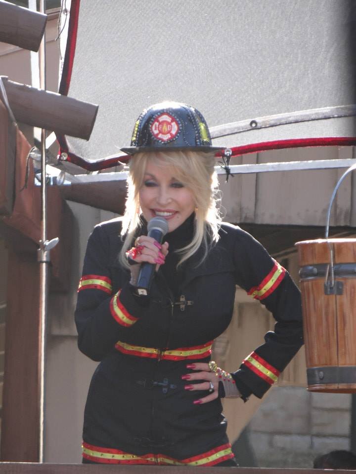 Dolly Parton stands at a microphone on an outdoor stage, wearing a navy firefighter's coat with reflective red and yellow stripes and a black fire helmet, one hand on her hip