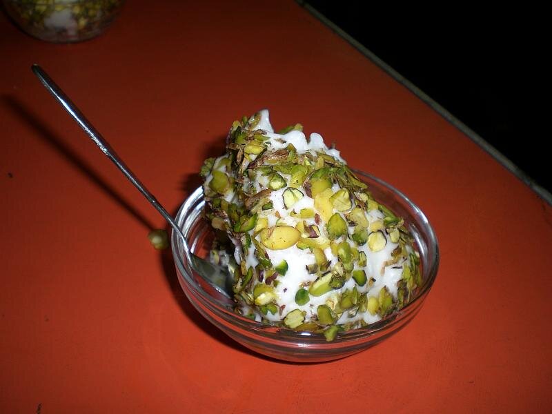 A glass bowl on an orange table holding a thick roll of white booza ice cream completely coated in coarsely chopped green pistachios, with a spoon resting in the bowl