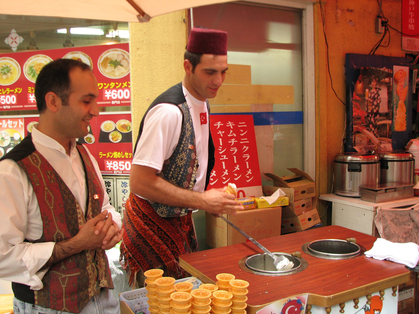 Two Turkish vendors in embroidered vests, one wearing a maroon fez, working a dondurma stall in Kyoto's Chinatown, with a long-handled paddle in a metal well and a stack of small waffle cones in the foreground