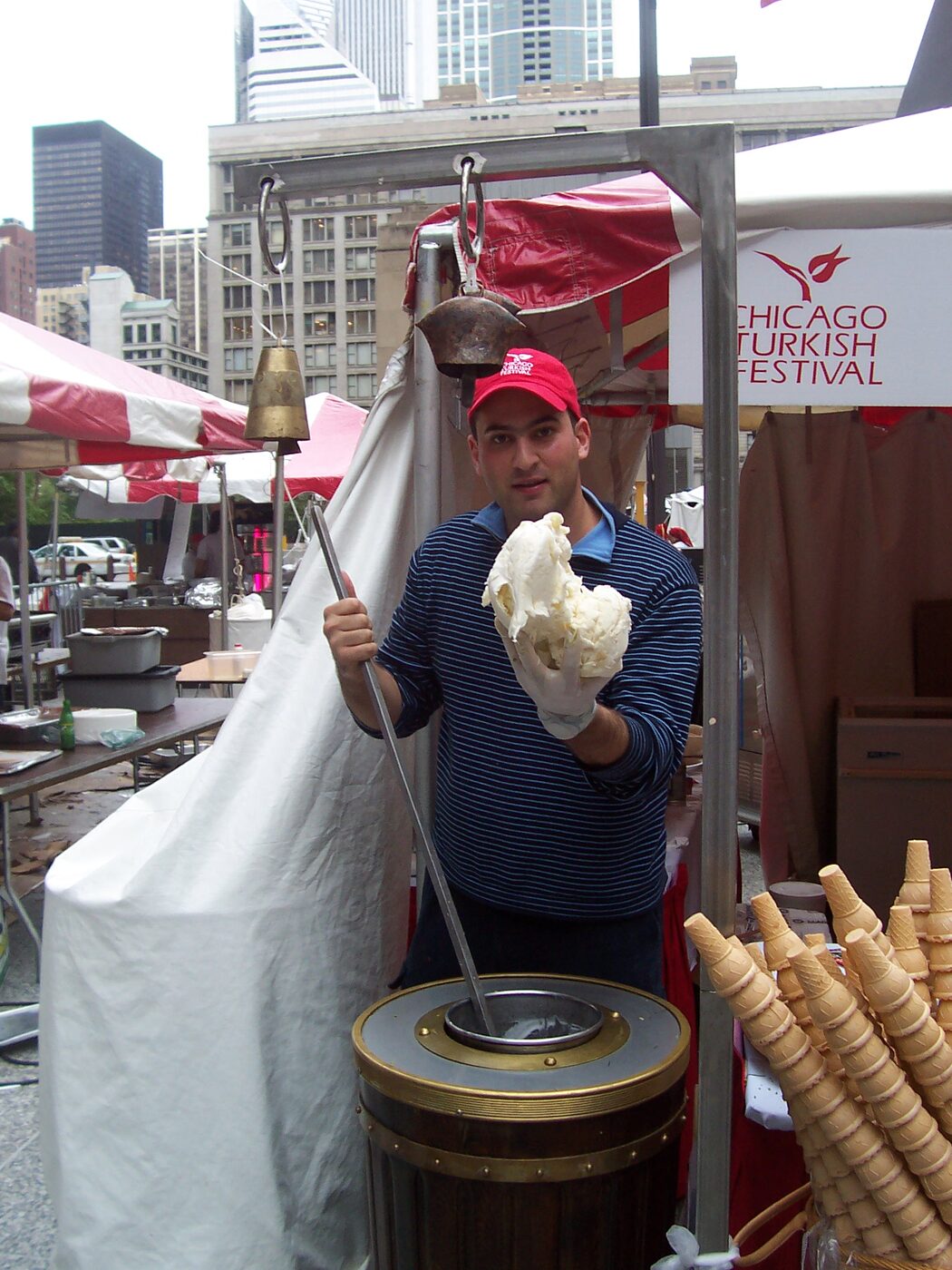 A vendor at a Turkish festival holding a pale, dough-like mass of dondurma in one gloved hand and a long-handled metal paddle in the other, with a wooden churn barrel in front of him and brass bells hanging overhead