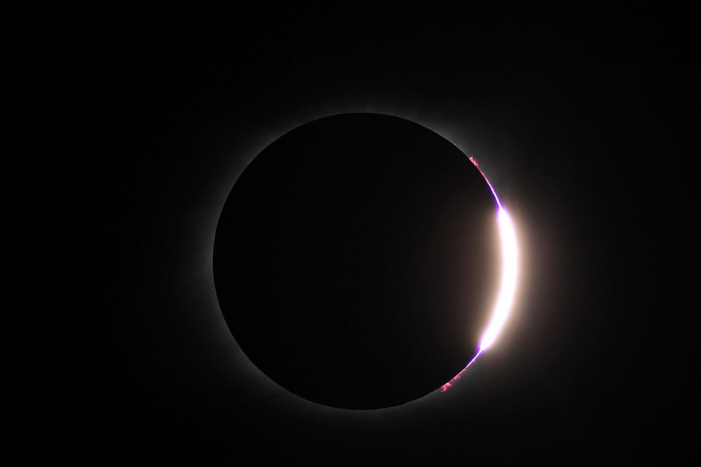 A dark lunar disc with a single brilliant white-gold burst of sunlight at its lower right edge, forming a ring of light around the darkness