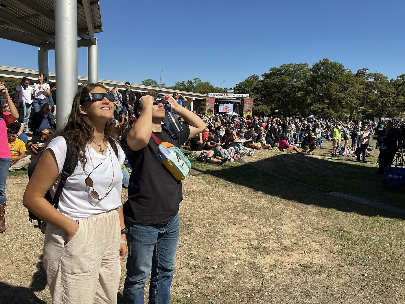 A crowd on a sunny lawn watching a solar eclipse, with two people in the foreground wearing cardboard eclipse glasses and looking up at the sky