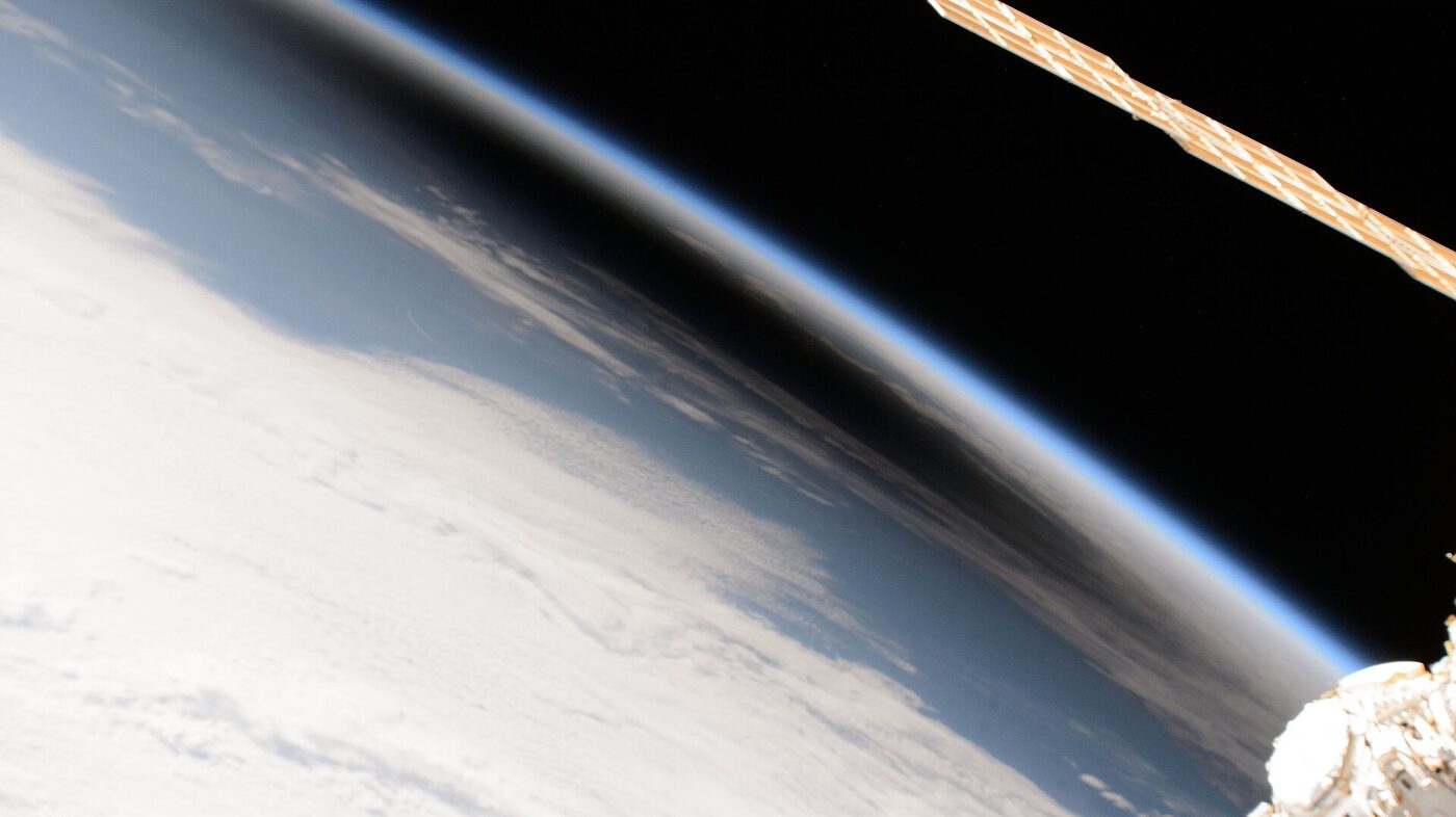 The curved edge of Earth photographed from the International Space Station, with a large dark patch of shadow lying across the cloud tops and the thin blue line of the atmosphere above