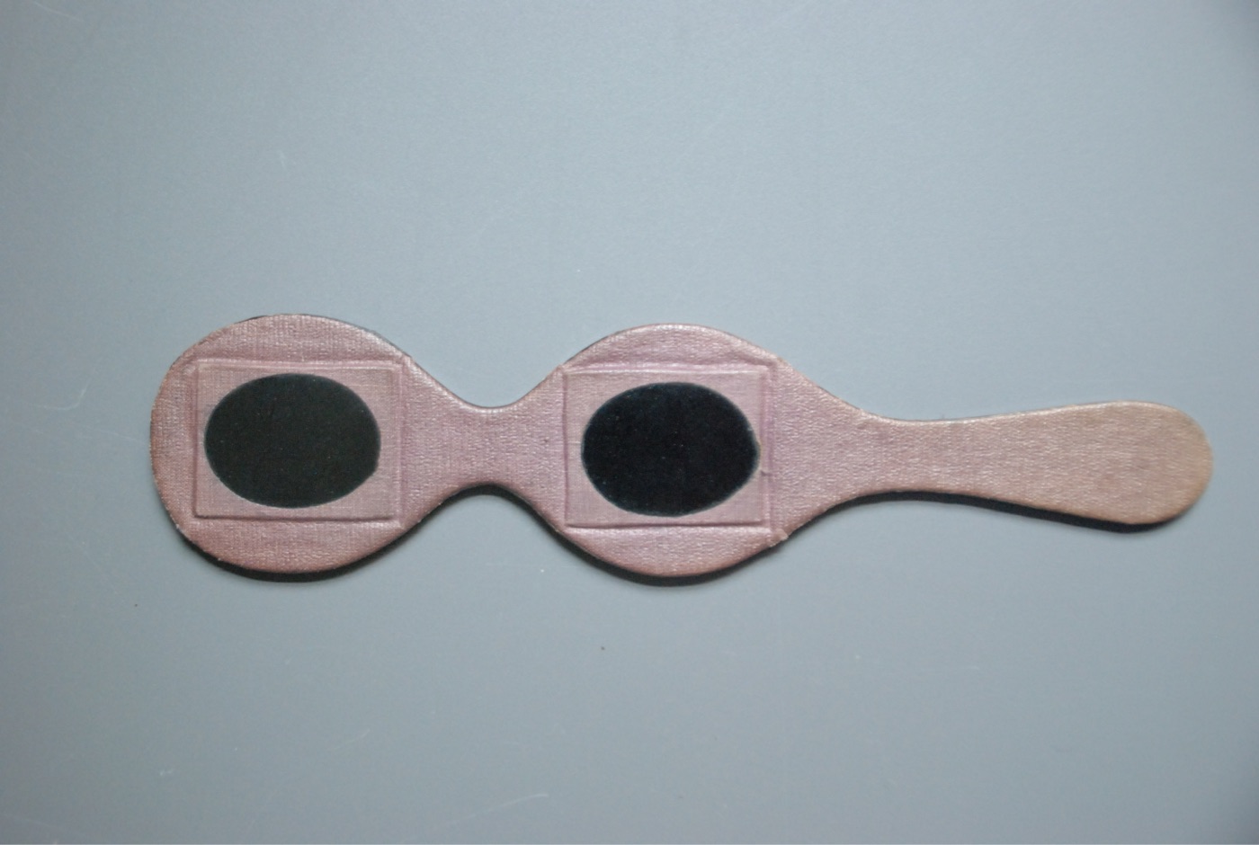 A pink hand-held eclipse viewer shaped like a lorgnette: two dark oval lenses in a shaped card body with a long handle to hold it up to the face