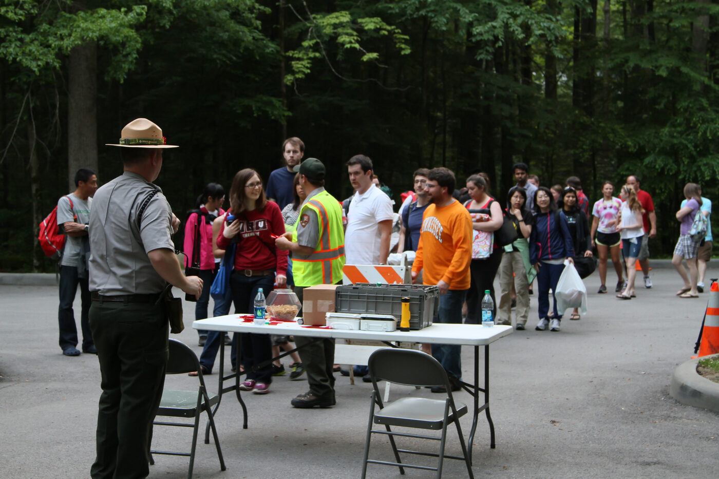 A park ranger in uniform standing at a folding table in a wooded parking area, with a crowd of casually dressed visitors of all ages gathered around waiting for the evening firefly shuttle