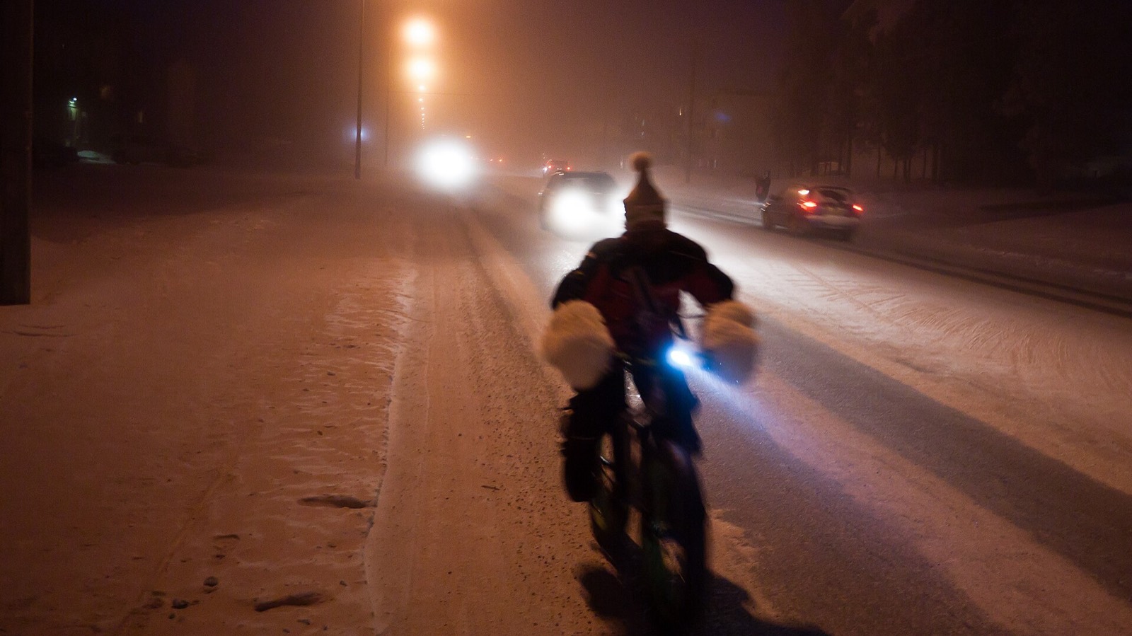 A cyclist riding through orange-lit ice fog on a snow-covered Whitehorse street at night, bundled in a parka, pogies on the handlebars, car headlights glowing through the haze behind