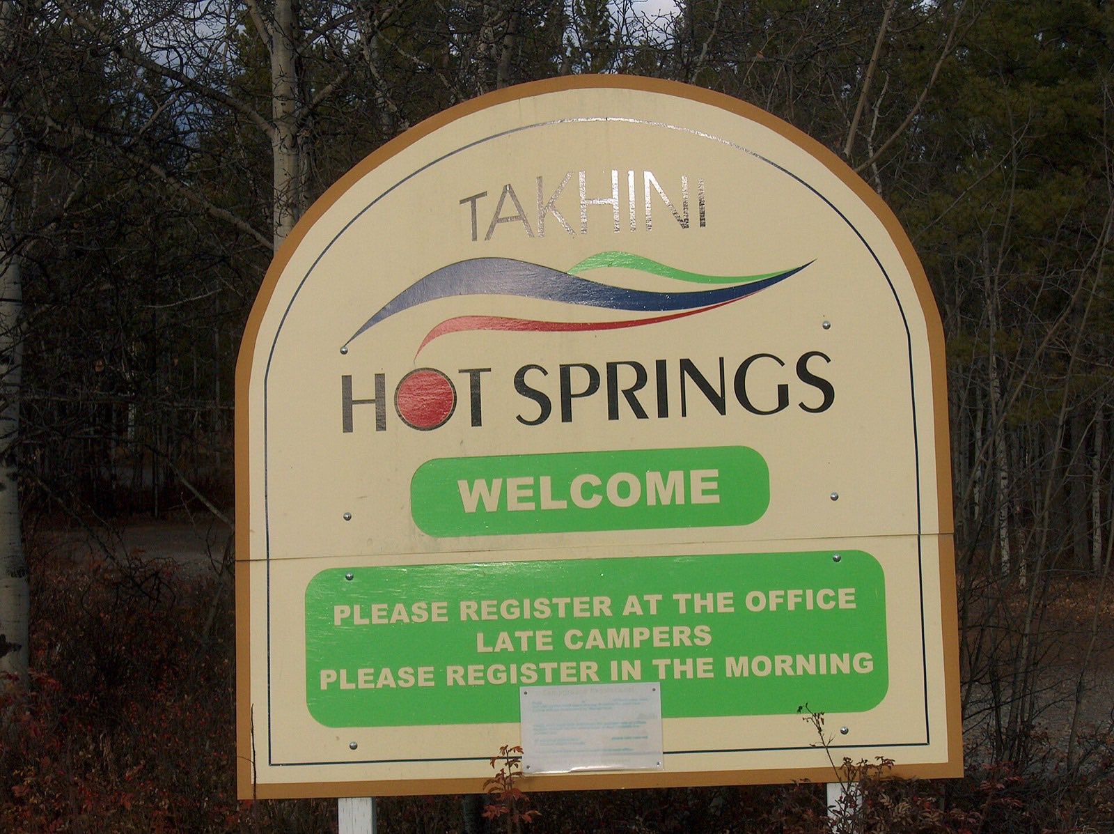 The Takhini Hot Springs welcome sign, a cream-coloured arched board with a red, blue and green swoosh logo, reading WELCOME and PLEASE REGISTER AT THE OFFICE, standing among bare birch trees