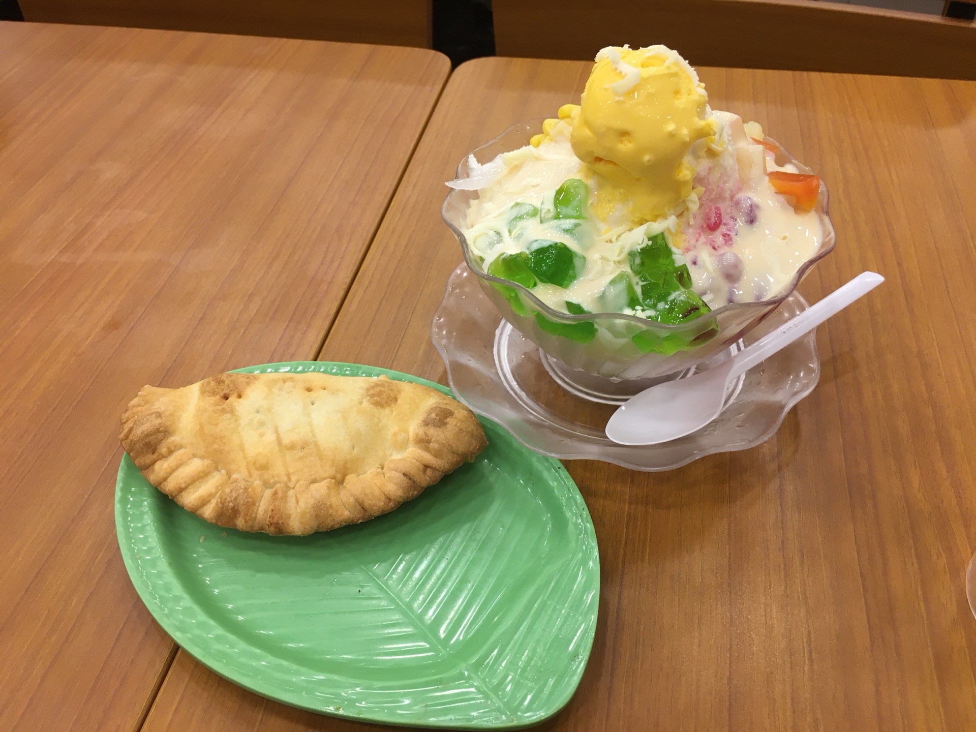 A golden baked empanada on a green leaf-shaped plate beside a fluted glass of halo-halo topped with ice cream and green jelly, on a wooden table