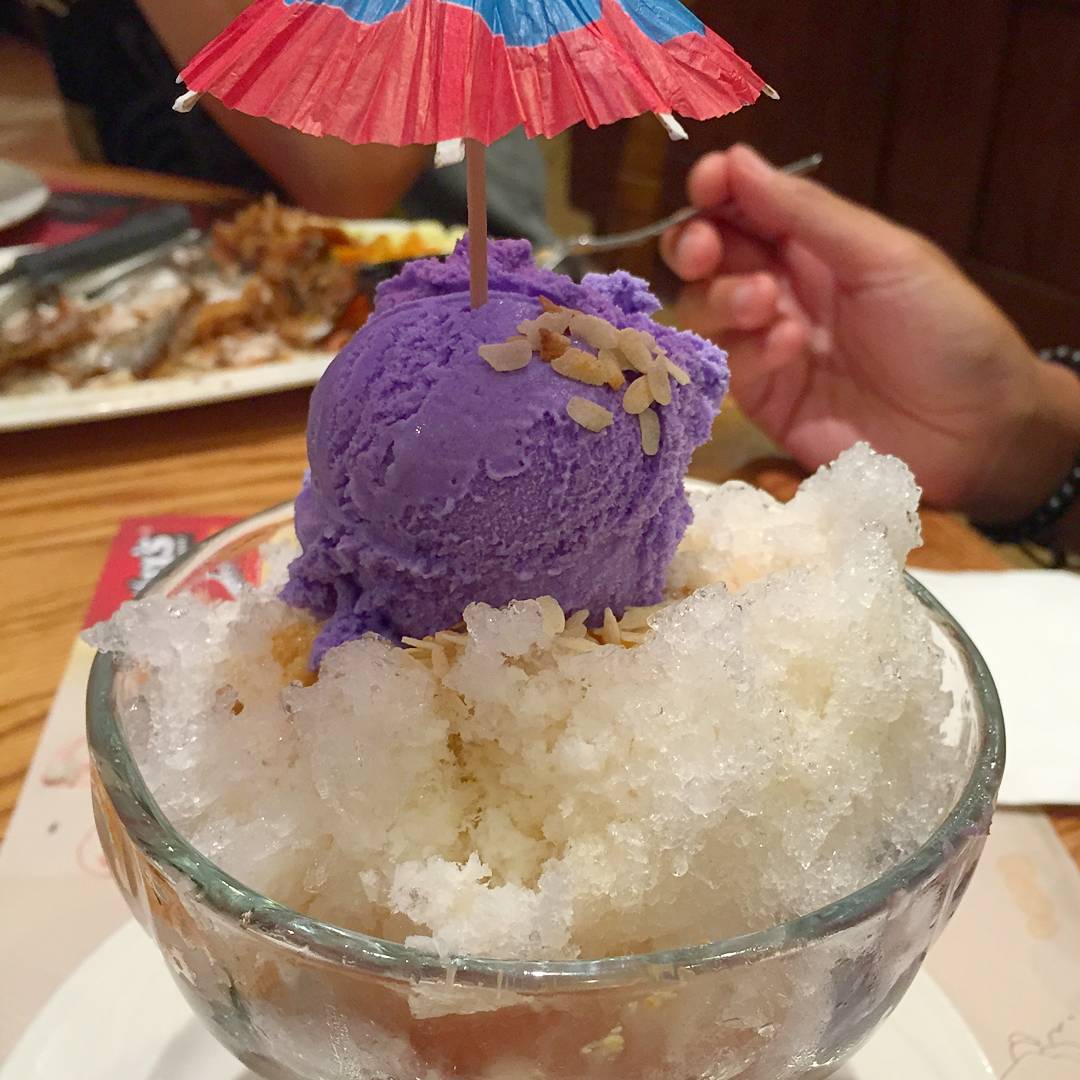 A glass bowl of halo-halo topped with purple ube ice cream, a little cocktail umbrella and a sprinkle of toasted rice flakes, held by a diner with a fork