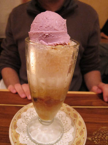A tall footed glass of halo-halo topped with a single scoop of purple ube ice cream, served on a paper-doily saucer