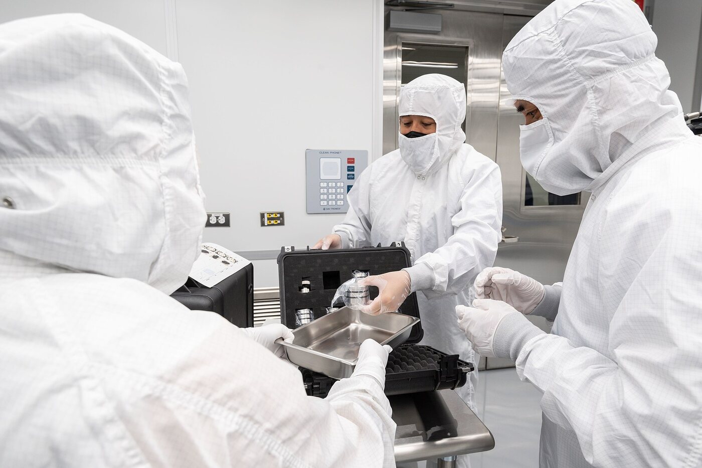 Three people in full white cleanroom bunny suits, hoods, masks and gloves standing around a black foam-lined transport case, lifting a small polished stainless steel canister out of it onto a metal tray