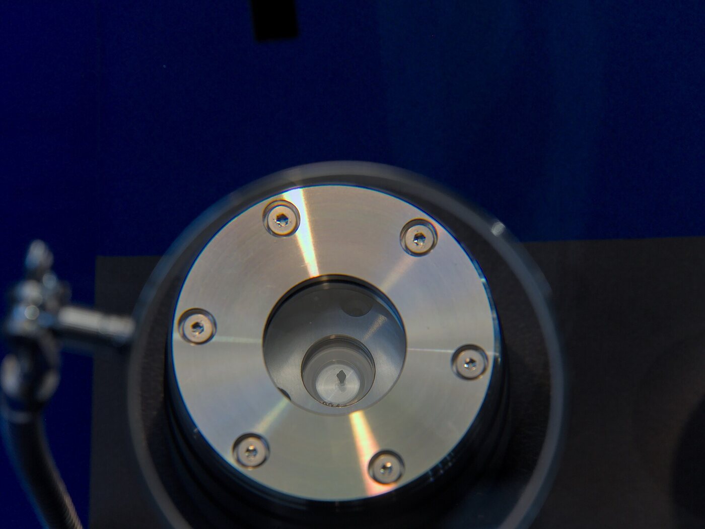 Close-up of a polished circular stainless steel container with six bolts around its rim and a small glass viewing port in the centre, through which a tiny dark speck is visible, photographed against a deep blue museum backdrop