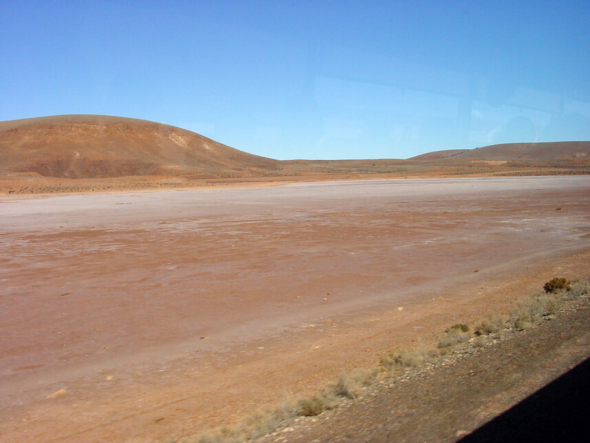 A wide, flat pale claypan of pinkish-orange dirt stretching to low bare hills under a cloudless deep blue sky, with a fringe of dry scrub in the foreground