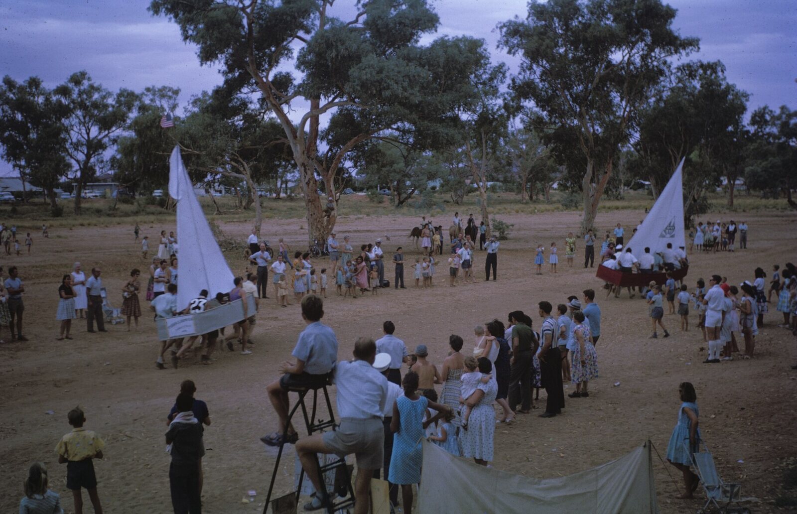 Faded colour slide from December 1962 showing two home-made boats with tall white sails being carried by running crews across the sandy Todd River bed, watched by a crowd in 1960s summer clothing, with gum trees, parked cars and horses in the background
