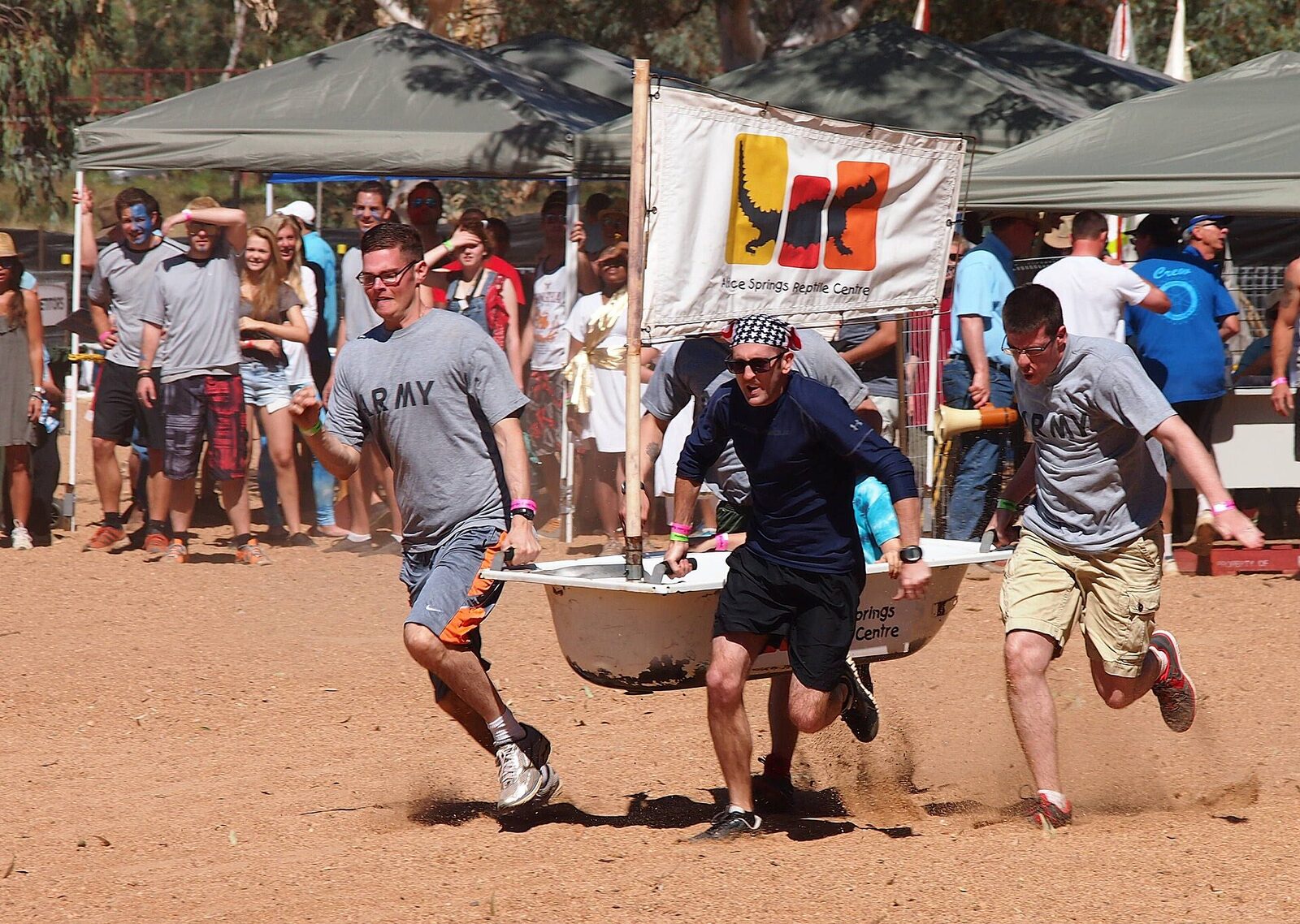 Three men in shorts and grey T-shirts sprint across the sand carrying a white bathtub fitted with a wooden mast and a sponsor's sail, kicking up dust, as a crowd of spectators cheers from under shade tents