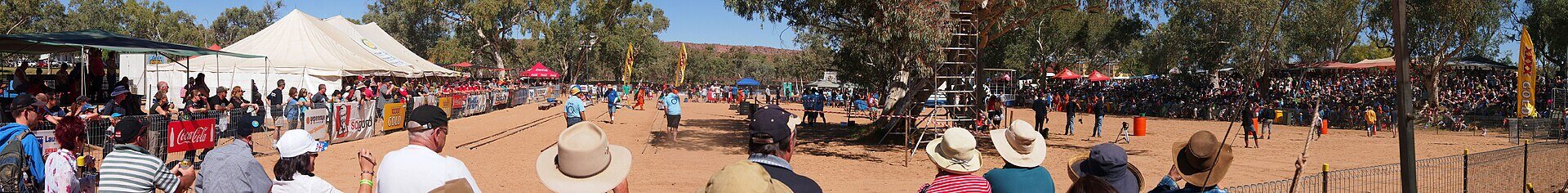 Wide panoramic view of the regatta course: a broad expanse of red sand lined on both sides by dense crowds under marquees and shade tents, with gum trees and the low red MacDonnell Ranges on the horizon