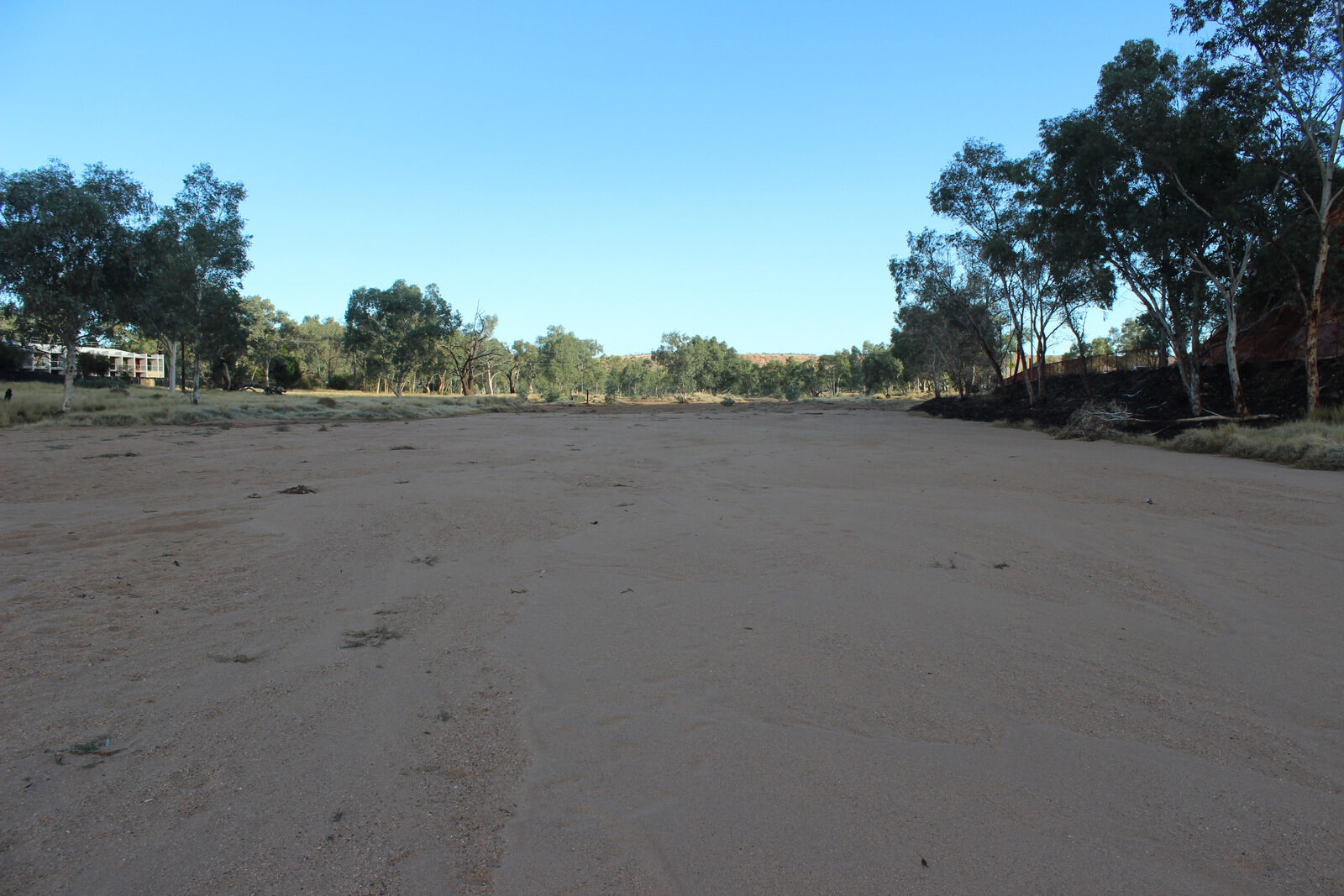 The empty bed of the Todd River at the Gap near Alice Springs: a wide flat channel of pale red sand under a clear blue sky, lined with river red gums, with a red rock outcrop on the right
