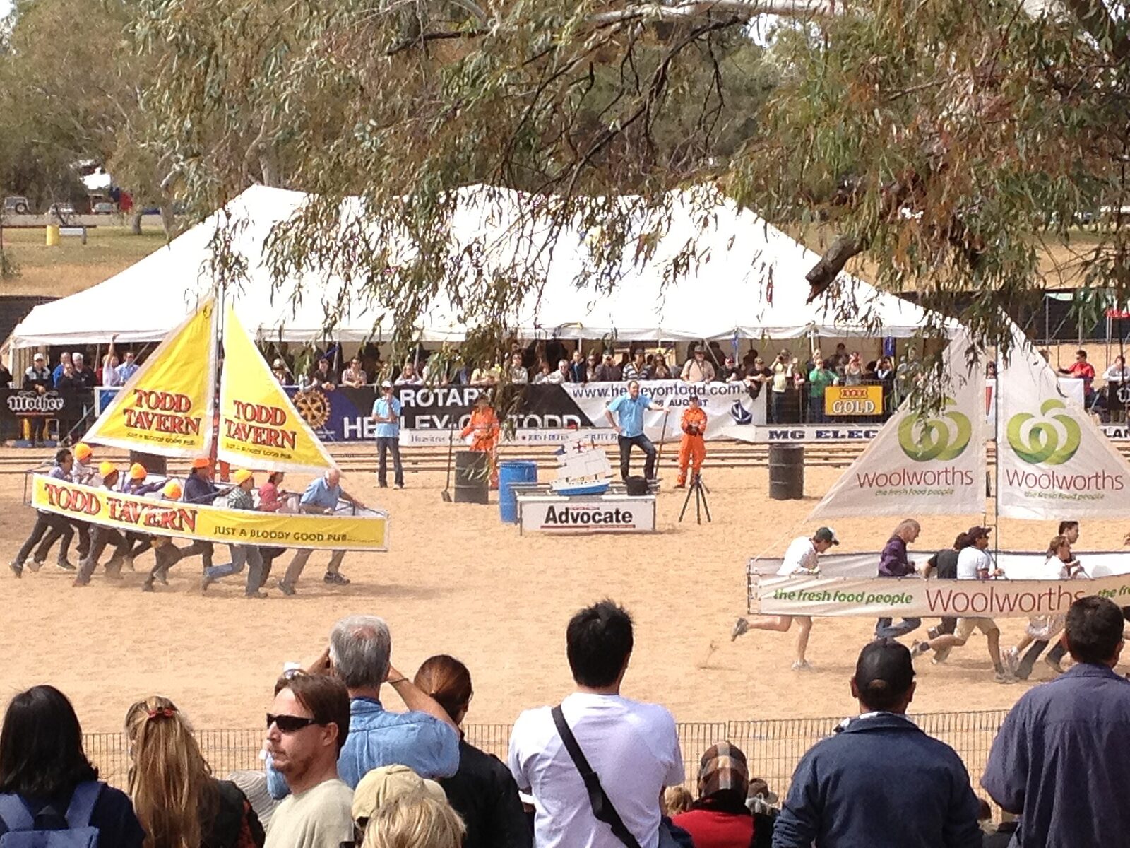 A crew in hard hats runs a bottomless yacht rigged with yellow Todd Tavern sails past the officials' area, where a banner reads ROTARY HENLEY-ON-TODD and www.henleyontodd.com.au, with a second crew running a Woolworths-branded boat on the right and a packed marquee of spectators behind