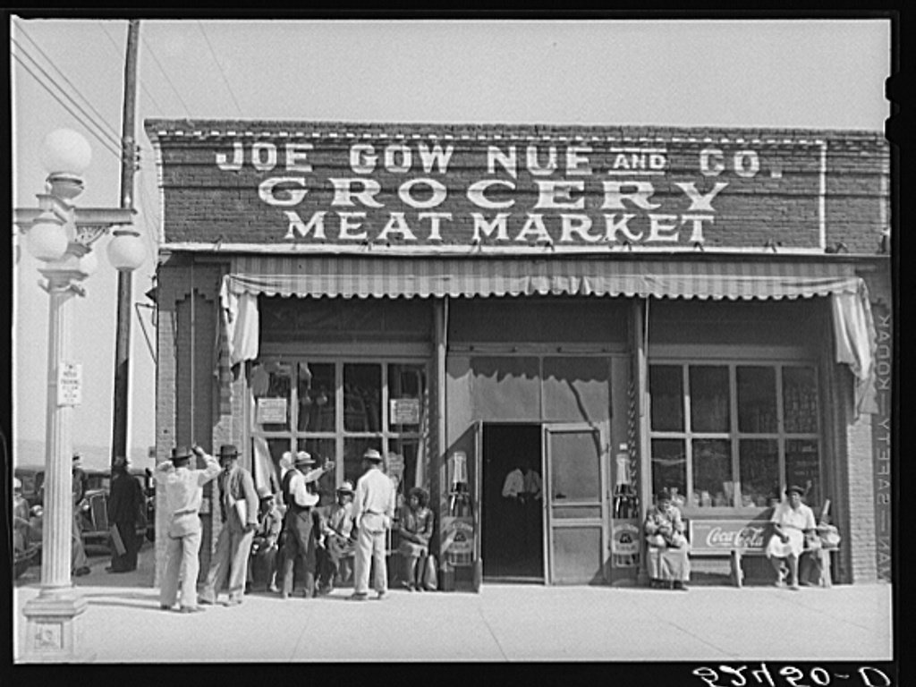 Black and white 1939 photograph of a brick storefront reading JOE GOW NUE AND CO. GROCERY MEAT MARKET, with a crowd of people standing and sitting on the sidewalk out front beneath Coca-Cola signs