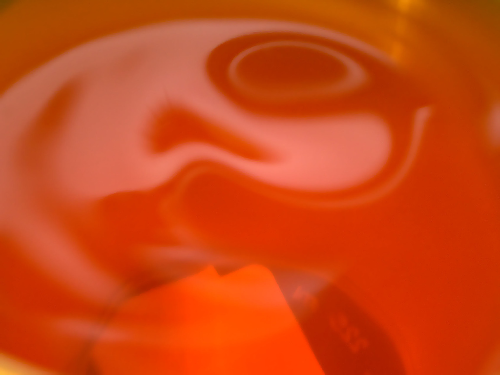 Extreme close-up of bright orange-red Kool-Aid in a glass pitcher, with a curl of liquid swirling on the surface