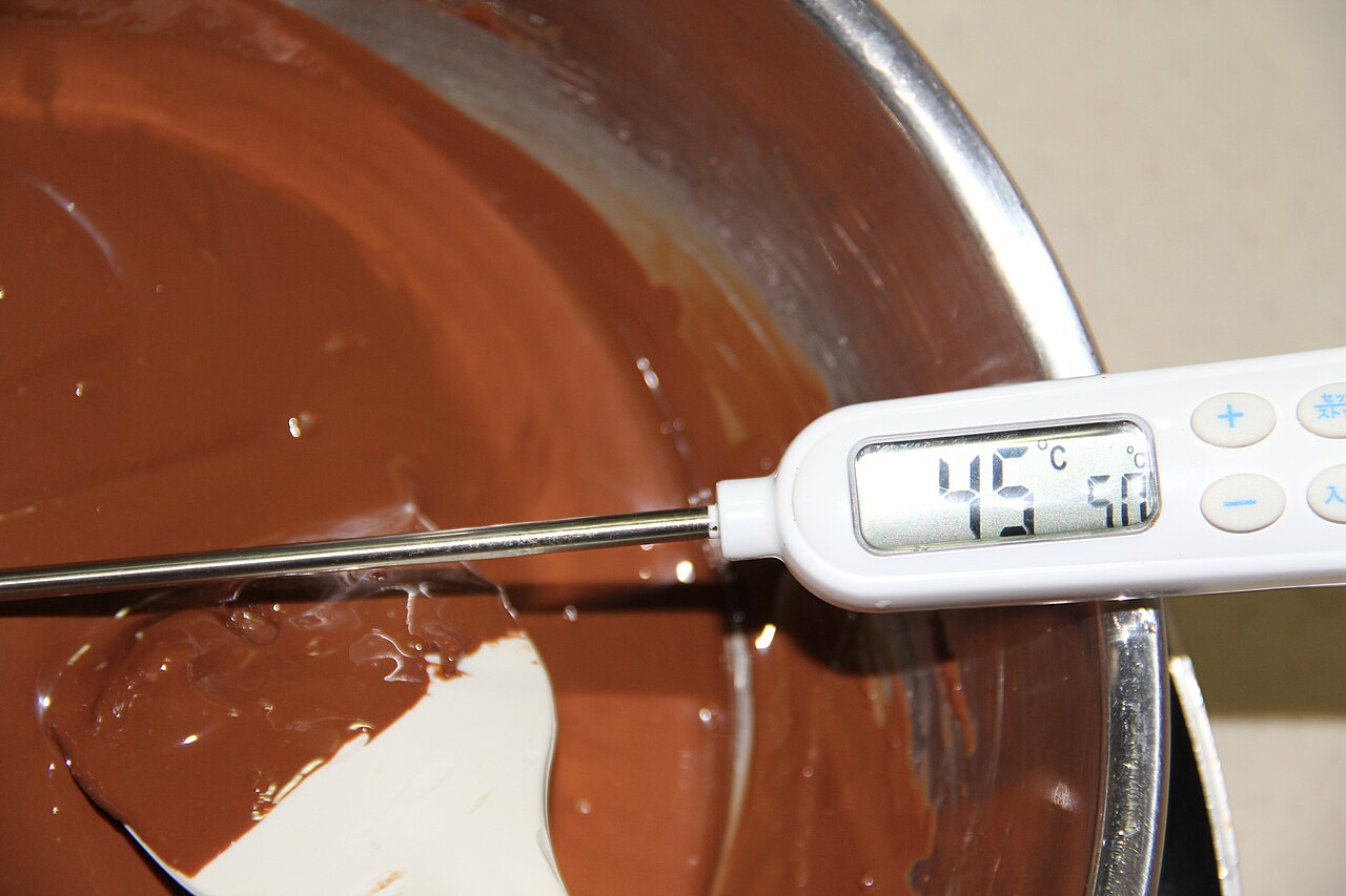 A stainless steel bowl of melted chocolate with a digital thermometer reading 45 degrees Celsius resting in it