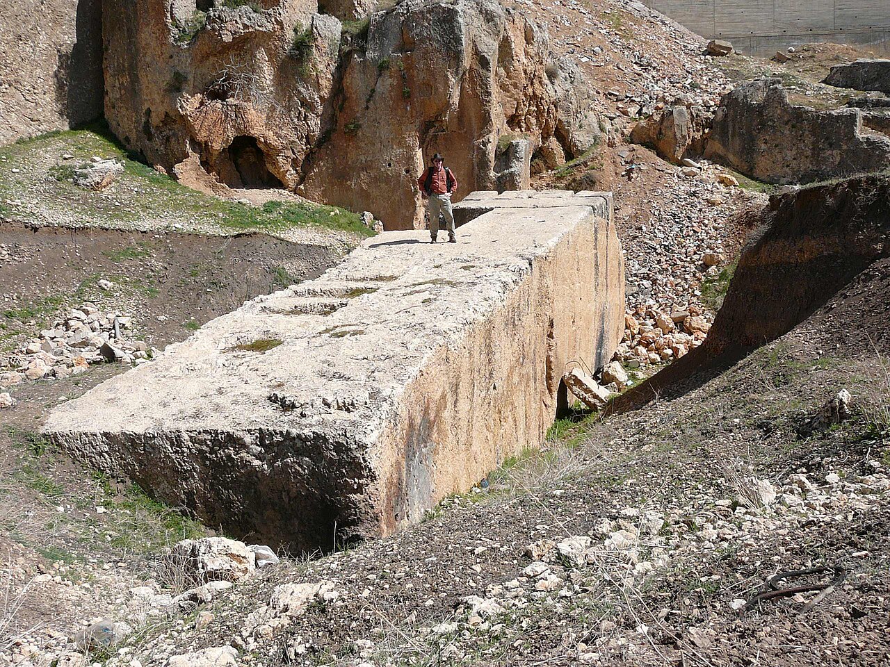 An enormous rectangular limestone block lying at a slight angle in a rocky quarry, its long flat top surface many times the length of a single adult figure standing on it near the far end, with excavated rubble slopes and green grass around its sides